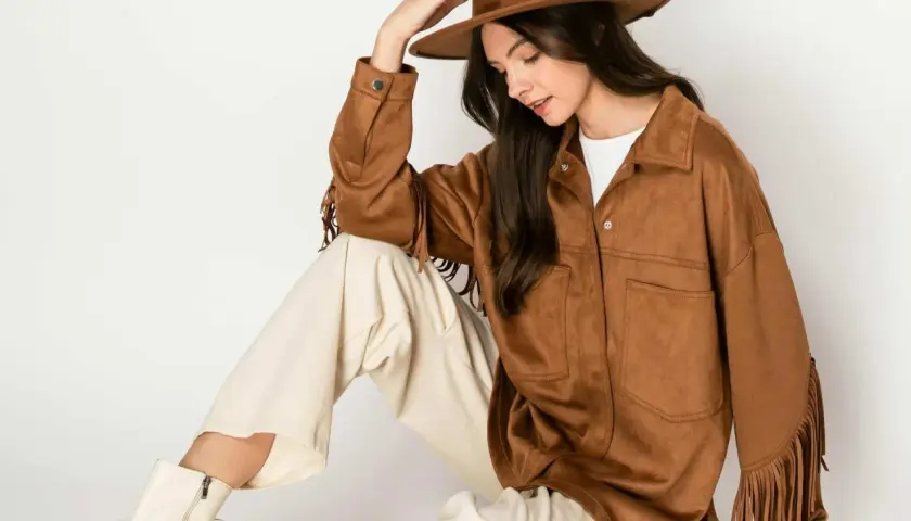 Mujer con sombrero y chaqueta de serraje que es suave y con flecos. Pantalones crema y botas blancas completan el look.