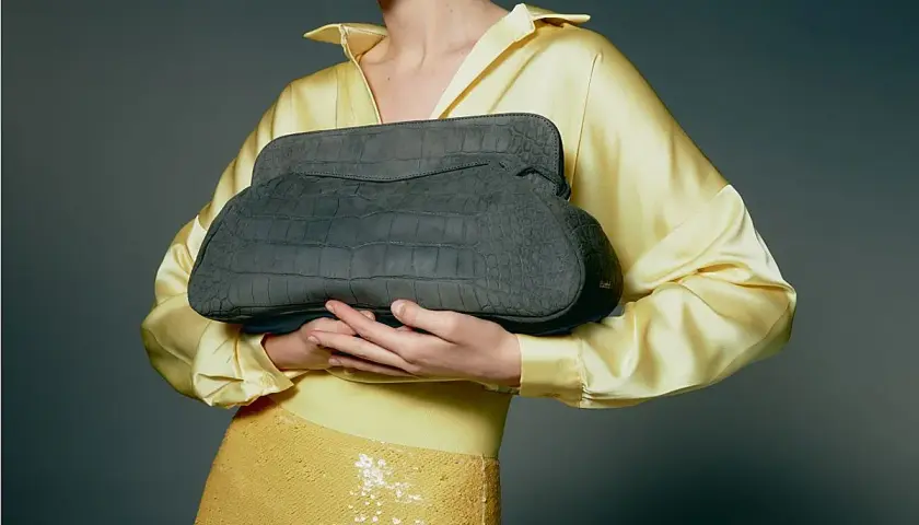 Elegante bolso gris de piel de cocodrilo, un accesorio de lujo de marcas españolas, sostenido por una mujer con blusa amarilla satinada y falda de lentejuelas.