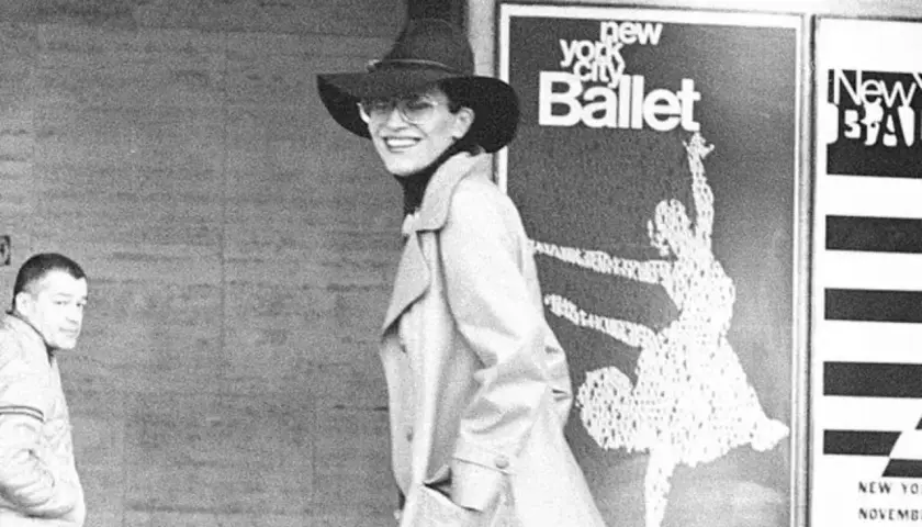 Elsa Peretti, con sombrero y gafas, sonríe frente a un cartel del New York City Ballet.