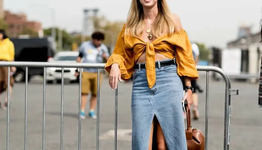 Mujer sonriente con un outfit falda vaquera larga, top amarillo anudado y bolso marrón.