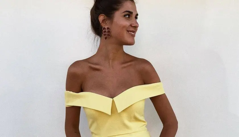 Mujer con moño alto y pendientes rojos, luciendo un vestido amarillo de hombros caídos. Ideas para combinar zapatos para vestido amarillo.
