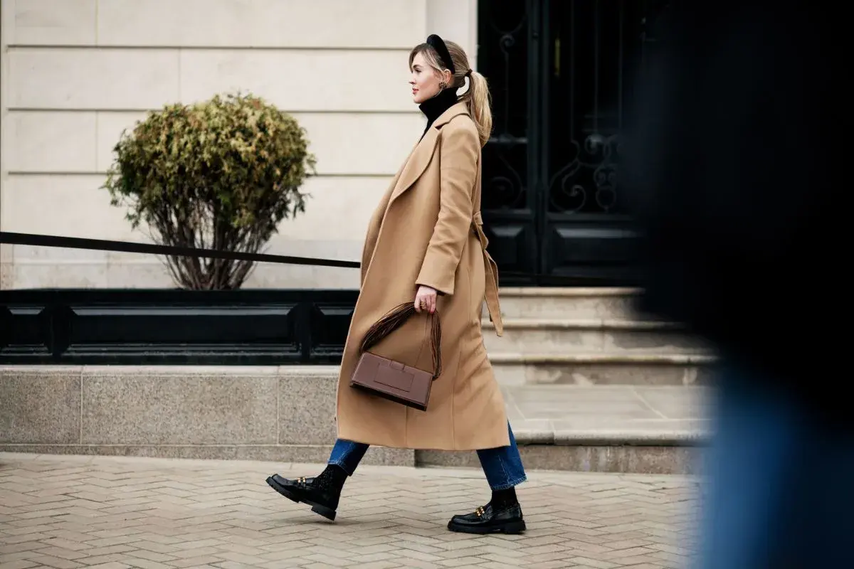 Mujer con abrigo camel, bolso marrón y mocasines negros, un look de quiet luxury paseando por la ciudad.