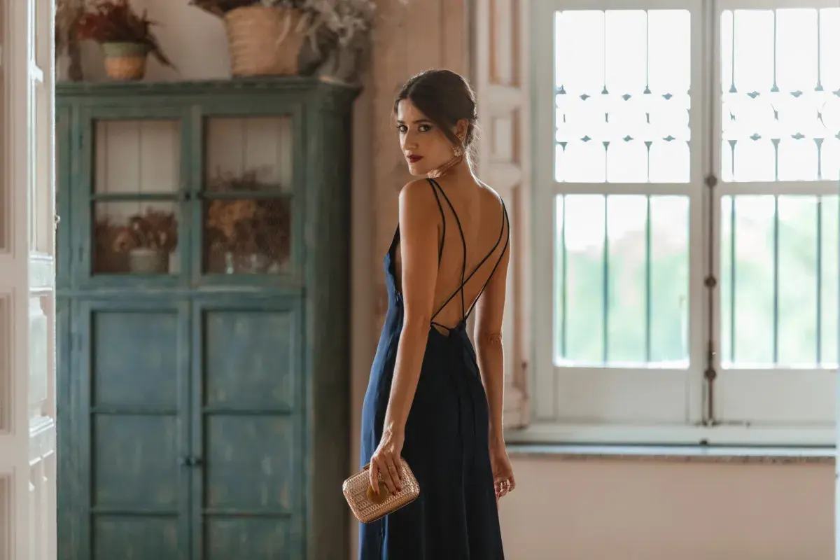 Mujer con un elegante vestido azul marino, perfecto para combinar en una boda. Lleva un bolso dorado y mira por encima del hombro.