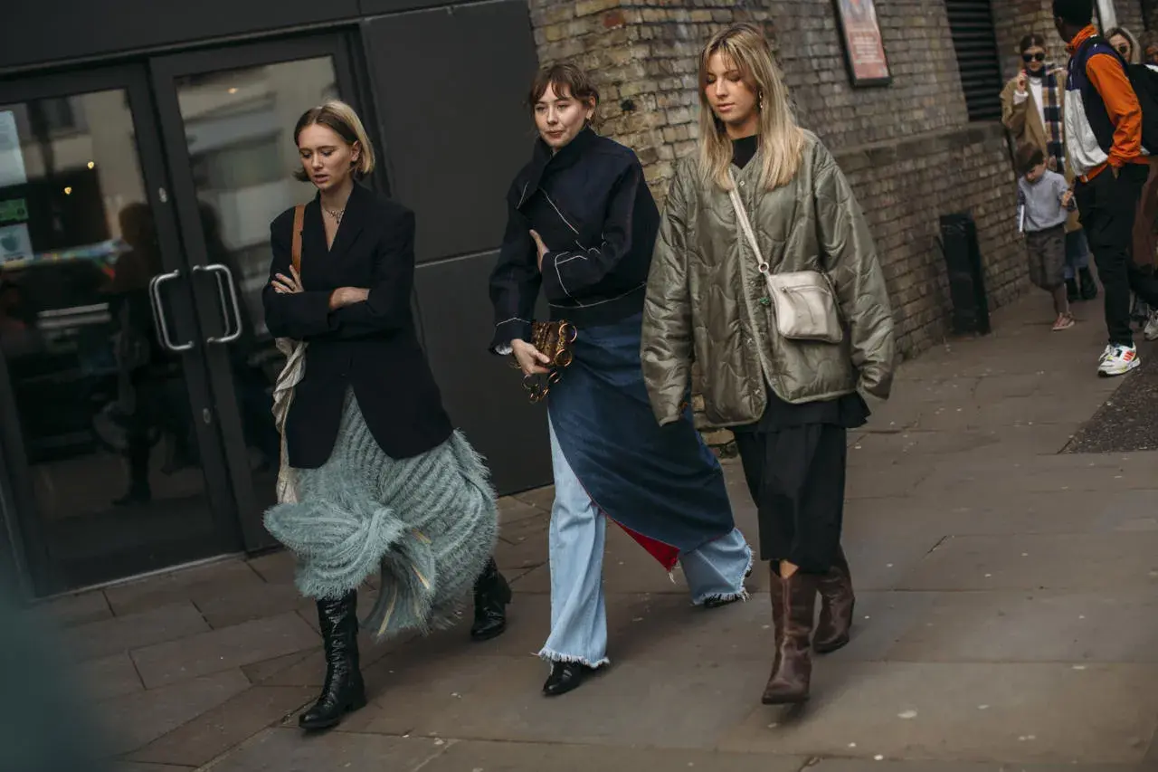 Tres mujeres caminan por la calle, cada una con un estilo único. La de la izquierda lleva unas **botas** negras altas y una falda de flecos. La del medio, un conjunto vaquero y unas botas negras. La de la derecha, unas botas marrones y una chaqueta aco...