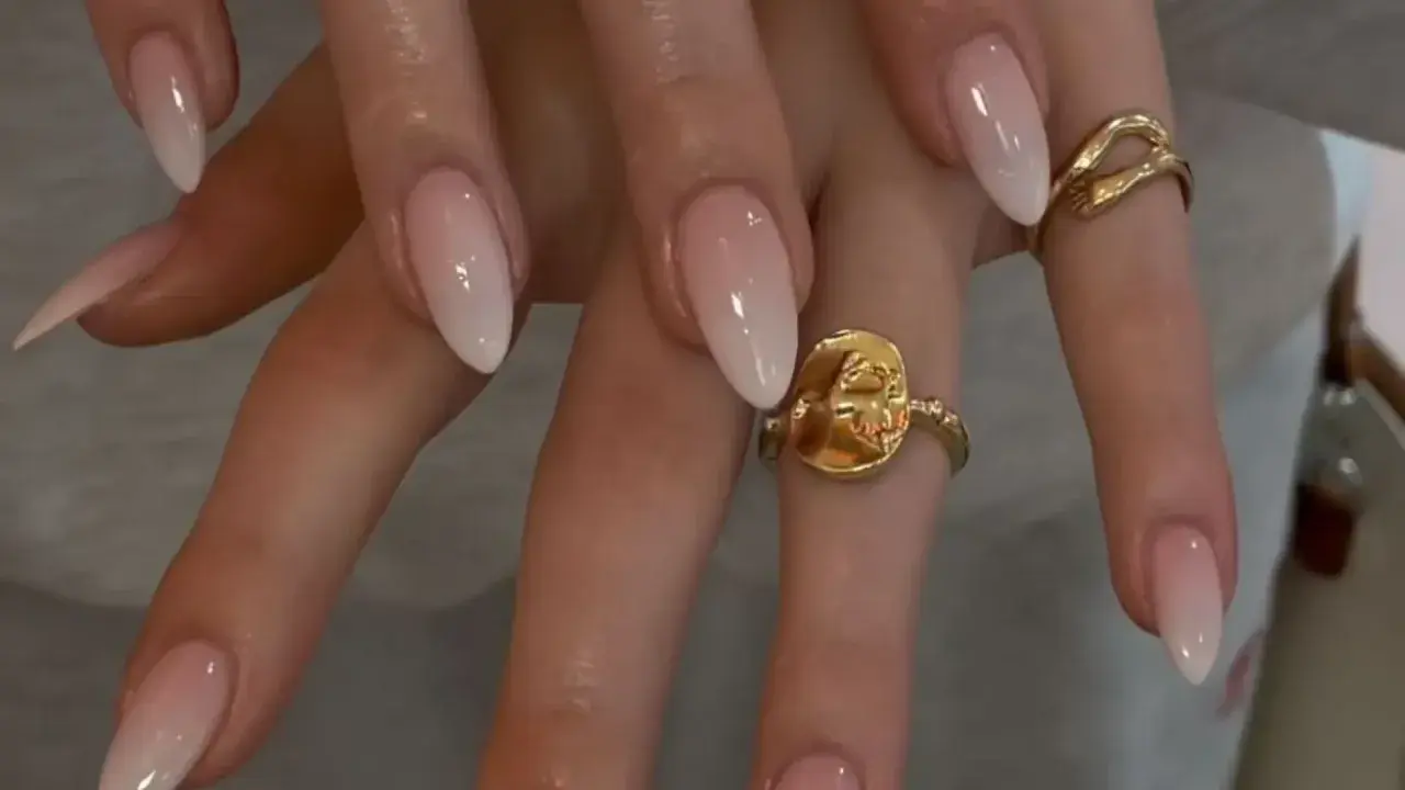Manos con uñas baby boomer natural, un degradado suave de rosa a blanco. Dos anillos dorados adornan los dedos.