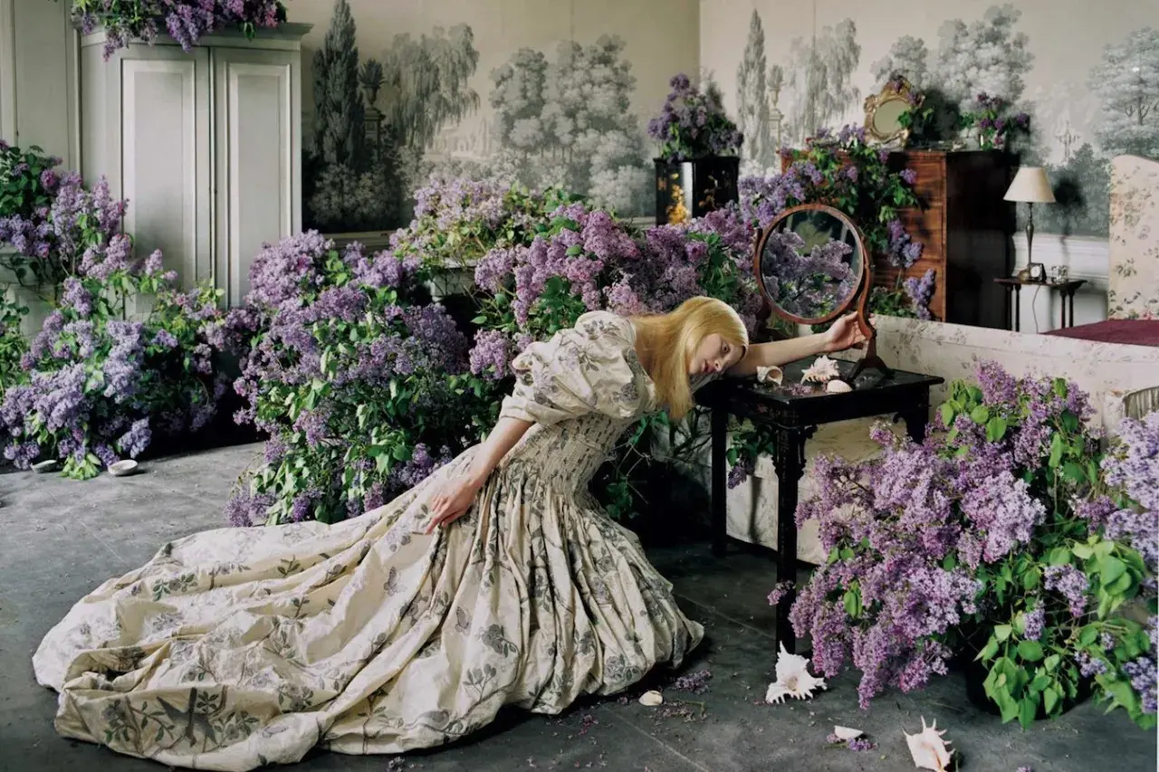 Una mujer rubia con un vestido estampado cae sobre un montón de lilas, evocando la estética onírica de Tim Walker.