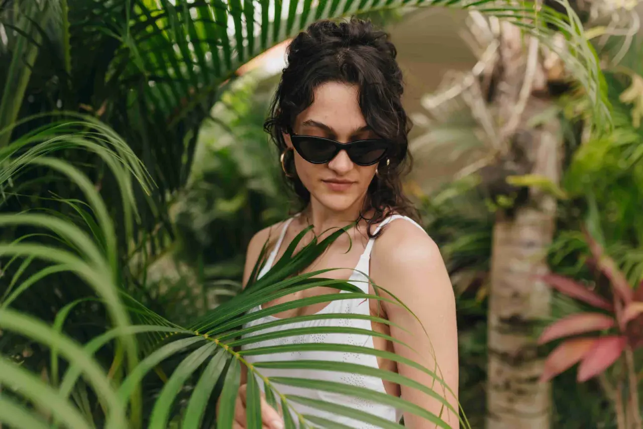 Joven con gafas de sol y traje de baño blanco entre hojas verdes, un ejemplo de moda ecológica.