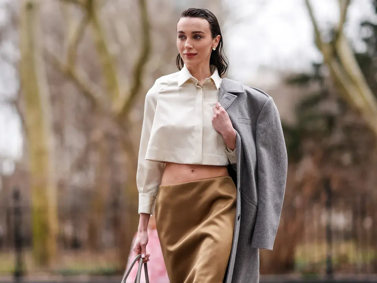 Mujer con blusa corta y falda satinada color caramelo, abrigo gris colgado del hombro.