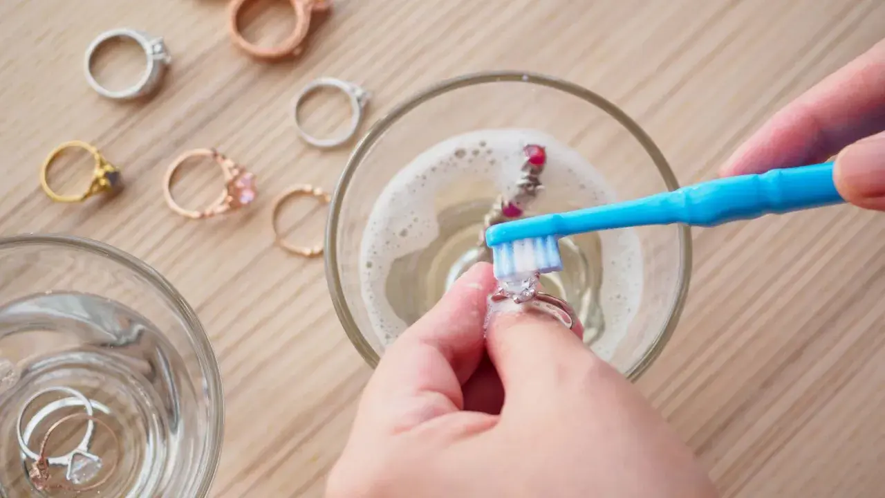 Manos limpian joyas con un cepillo azul en agua con espuma. Otros anillos esperan su turno.