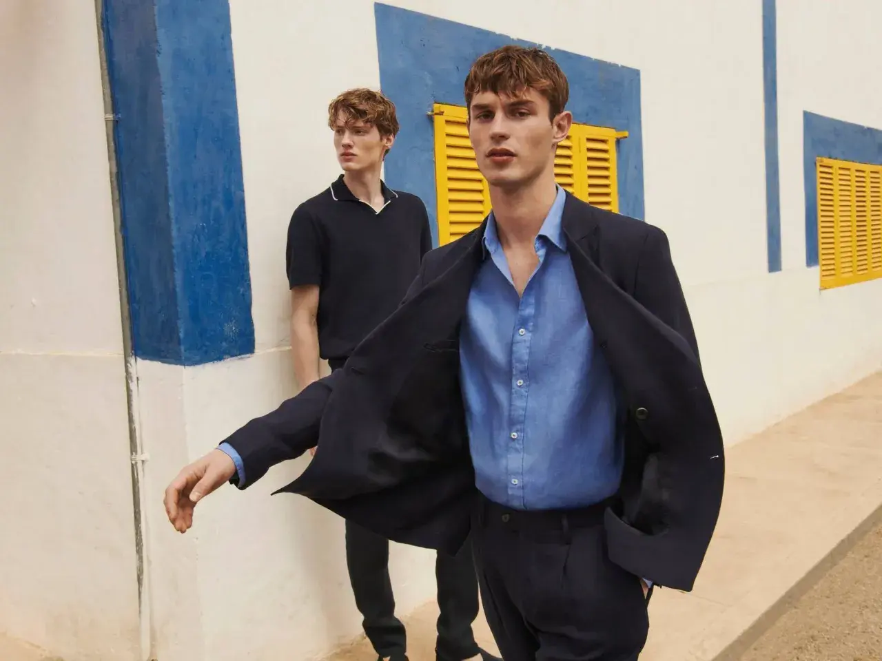 Dos hombres modelan un outfit oficina verano hombre: uno con chaqueta azul marino y camisa de lino, otro con polo oscuro.