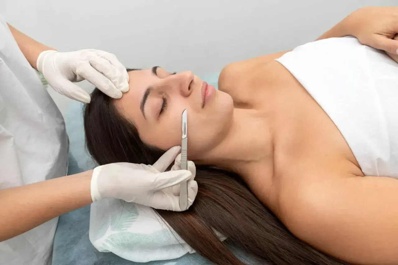 Una esteticista realiza un dermaplaning facial a una mujer, eliminando vello fino y células muertas con un bisturí.