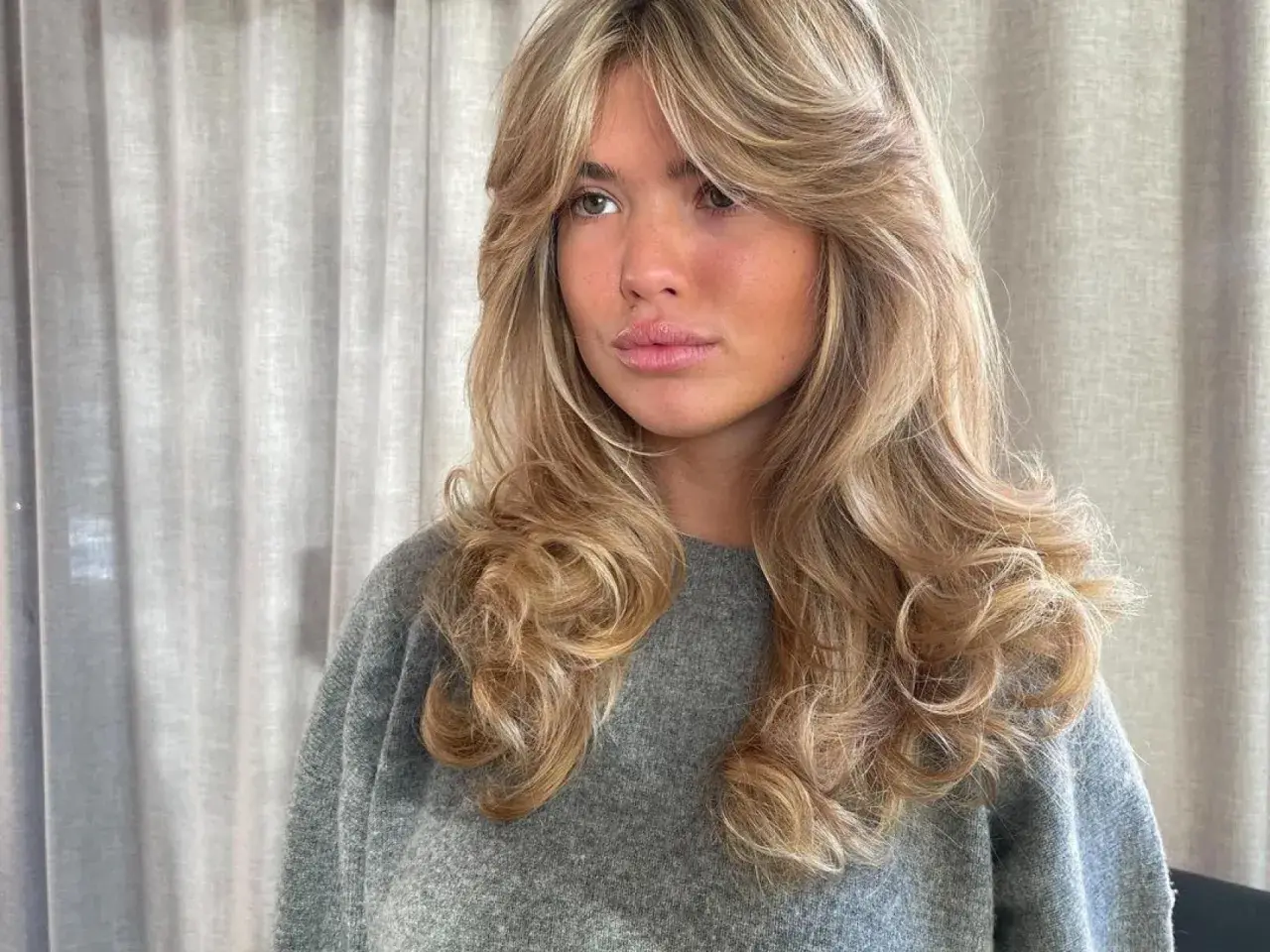 Mujer con cabello rubio y ondas voluminosas, un corte **hush cut** que enmarca su rostro.