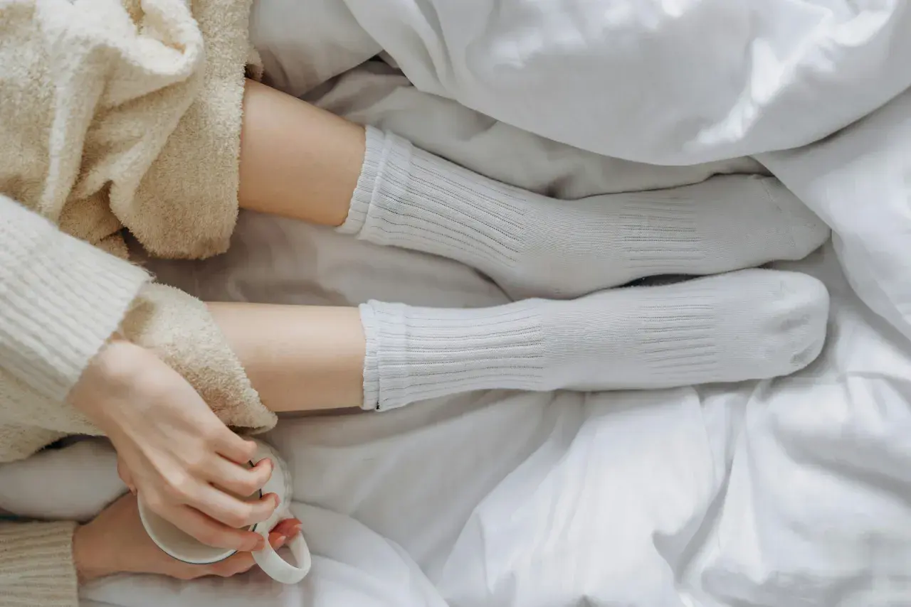 Piernas con calcetines blancos de poliamida, envueltas en mantas suaves, sosteniendo una taza.