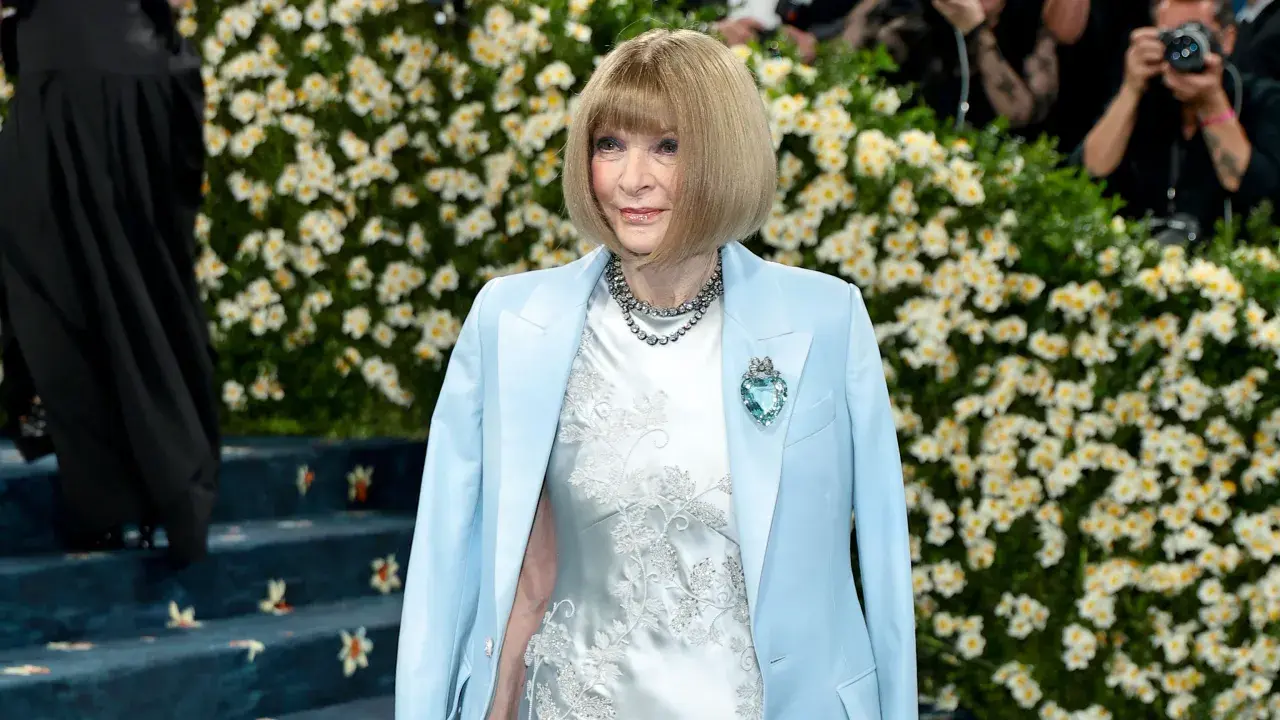 Anna Wintour, figura clave de que es la Gala Met, posa con un traje azul claro y un vestido blanco bordado, adornada con un collar y un broche de corazón.