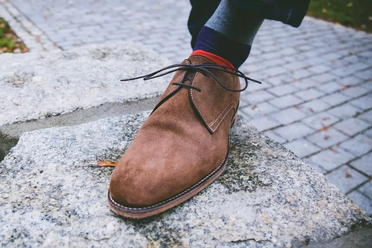 Zapato de gamuza marrón con cordones negros, calcetín a rayas y un consejo sobre como quitar manchas de humedad en zapatos de piel.