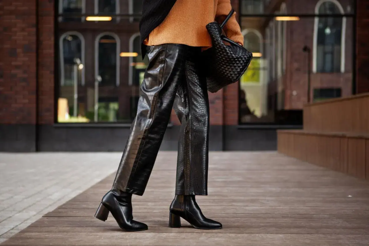 Elegante outfit con botas altas negras, pantalones de cuero texturizado y suéter naranja.