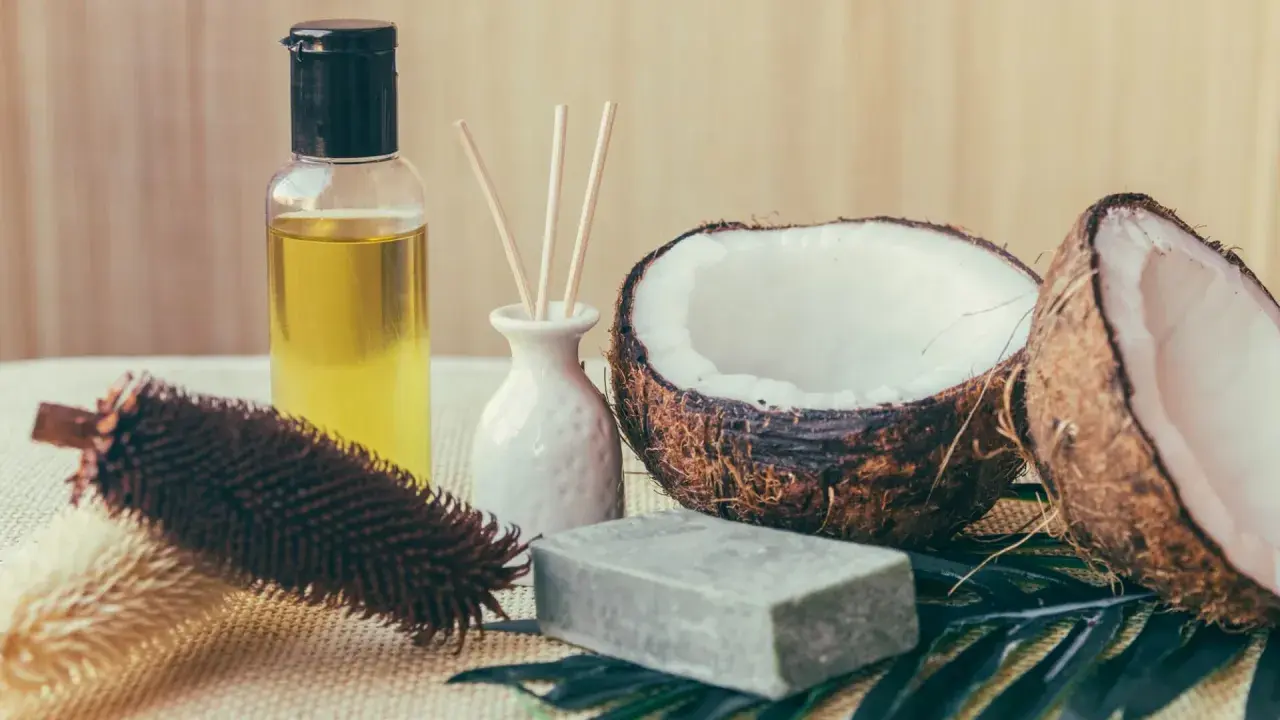 Aceite de coco para el pelo, junto a un coco partido, jabón y un cepillo.