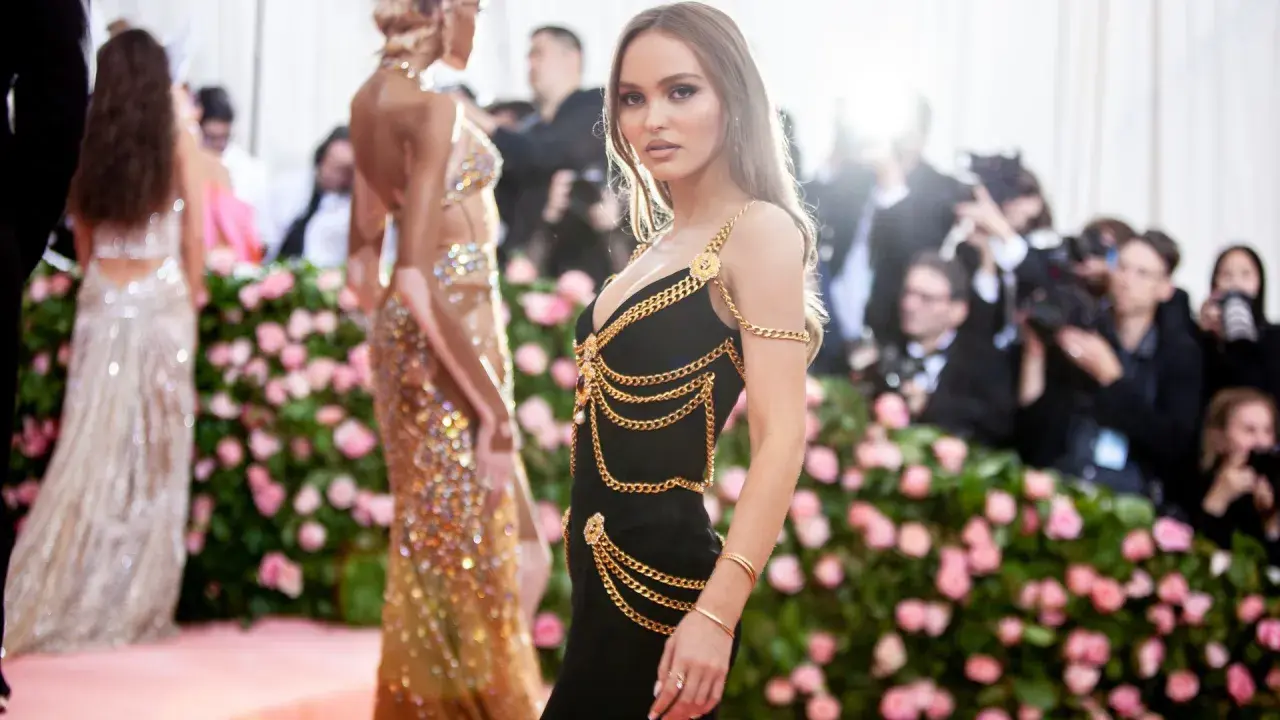 Lily-Rose Depp deslumbrante con un vestido negro adornado con cadenas doradas, posando ante un fondo de flores rosas y fotógrafos.