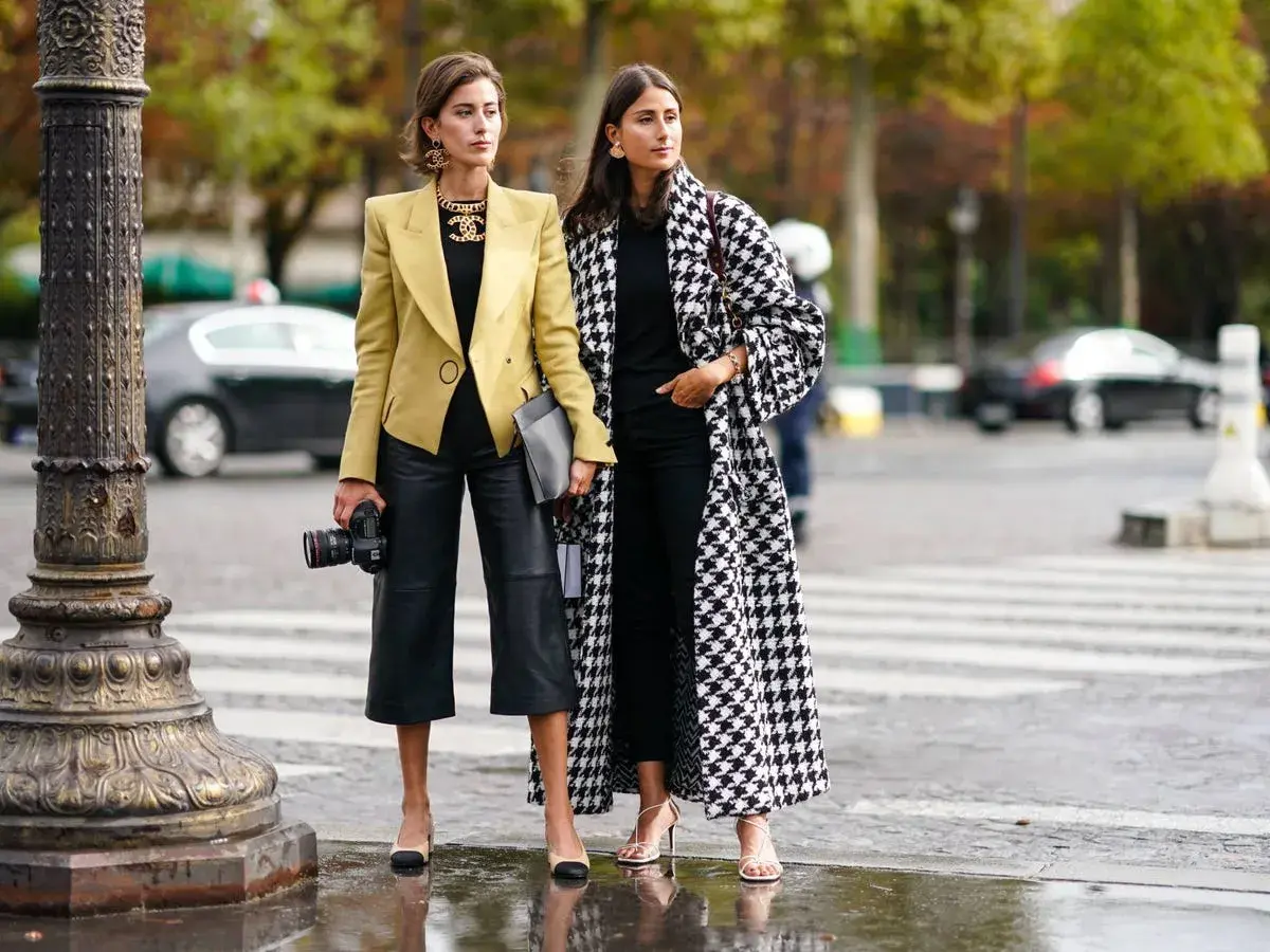 Dos mujeres con estilo muestran cómo vestir en otoño en España: una con blazer amarillo y pantalón culotte de cuero, la otra con abrigo de pata de gallo.