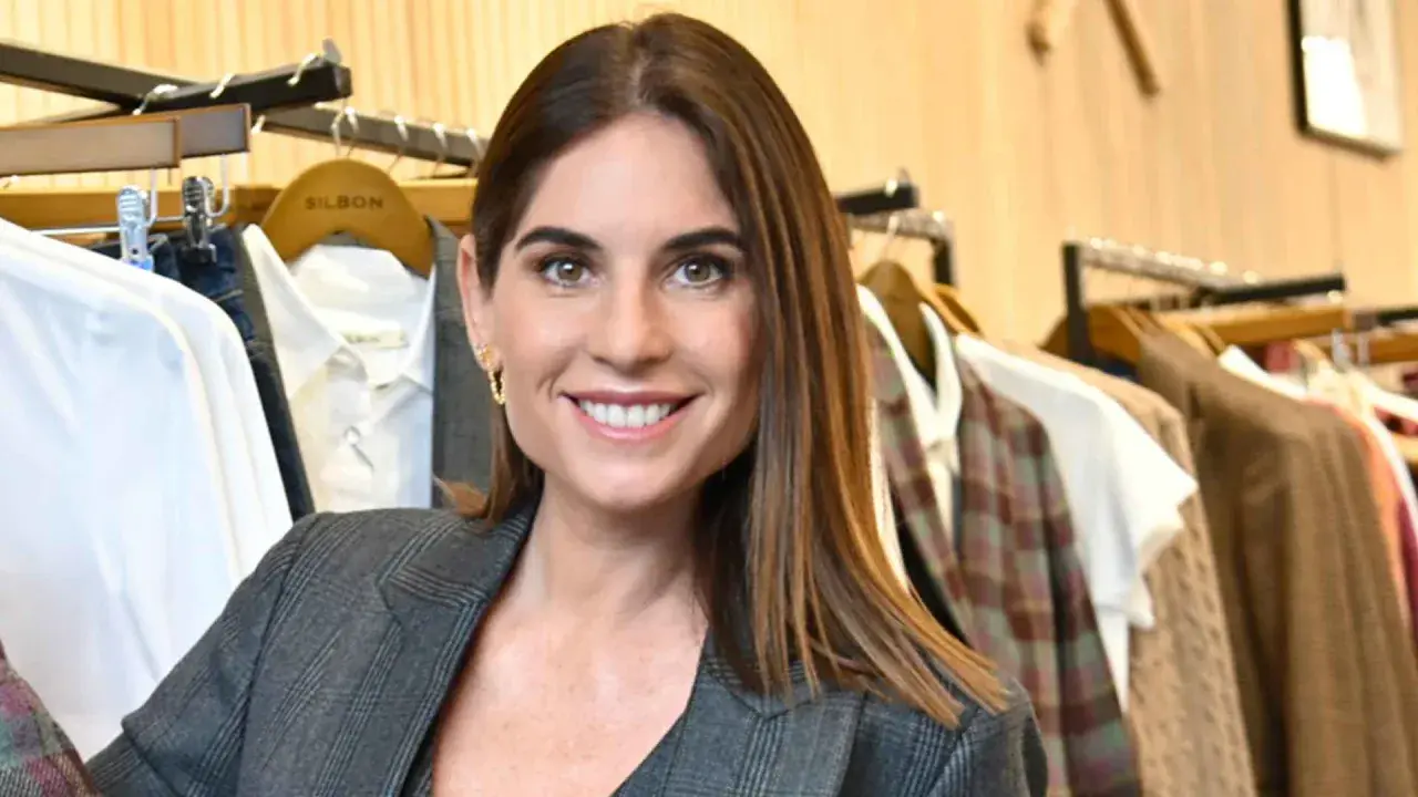 Lourdes Montes sonríe con un traje gris a cuadros, rodeada de ropa colgada en perchas en una tienda.