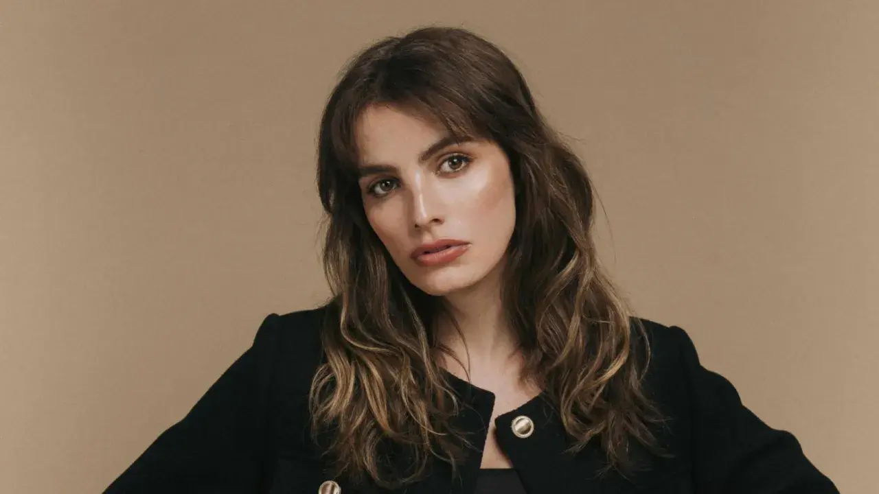 Retrato de una mujer con cabello castaño ondulado y flequillo, luciendo un blazer negro. Su mirada es intensa, transmitiendo un aire de misterio y elegancia, como si fuera la protagonista de una historia de lux pascal.