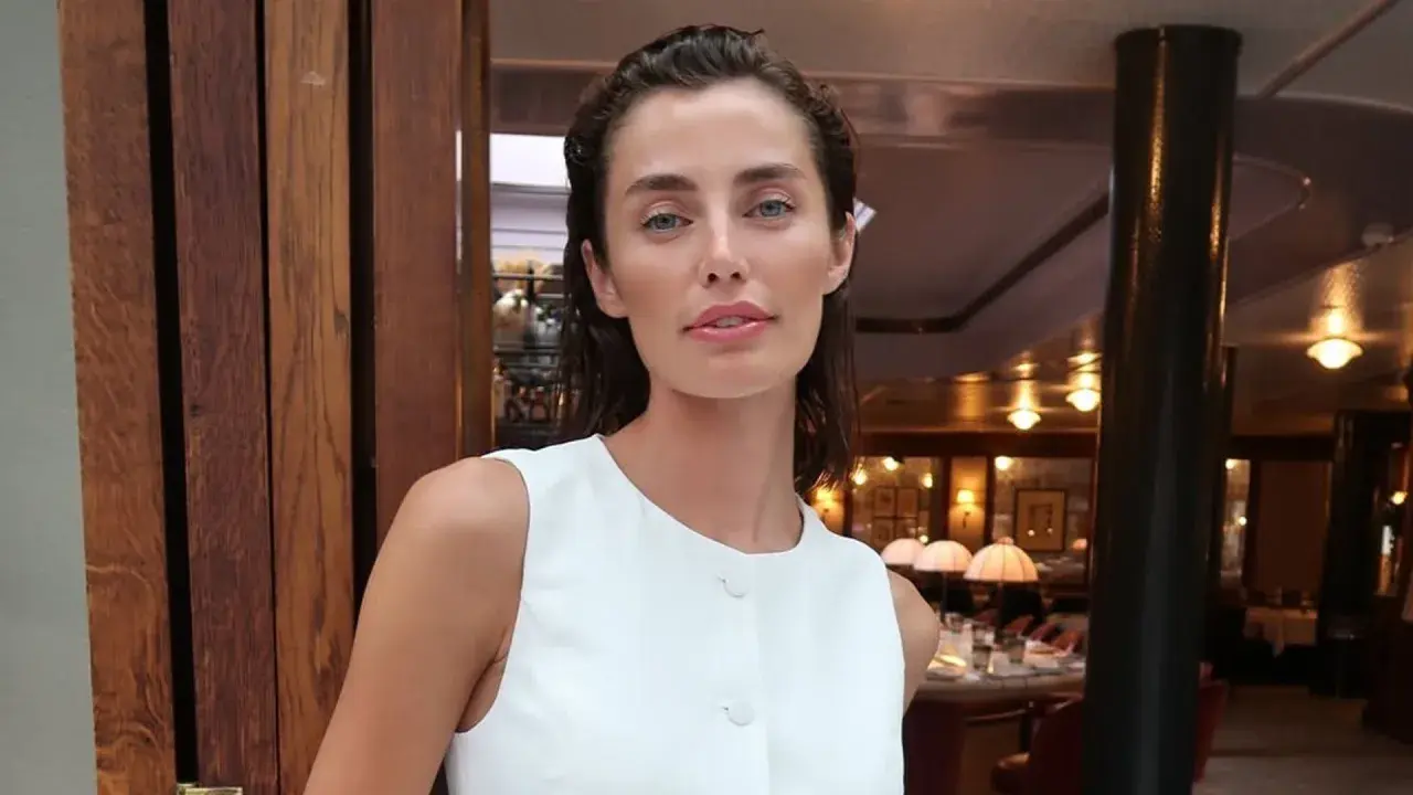 Rebecca Donaldson, con su mirada penetrante y labios rosados, posa con un elegante vestido blanco en un restaurante con ambiente cálido.