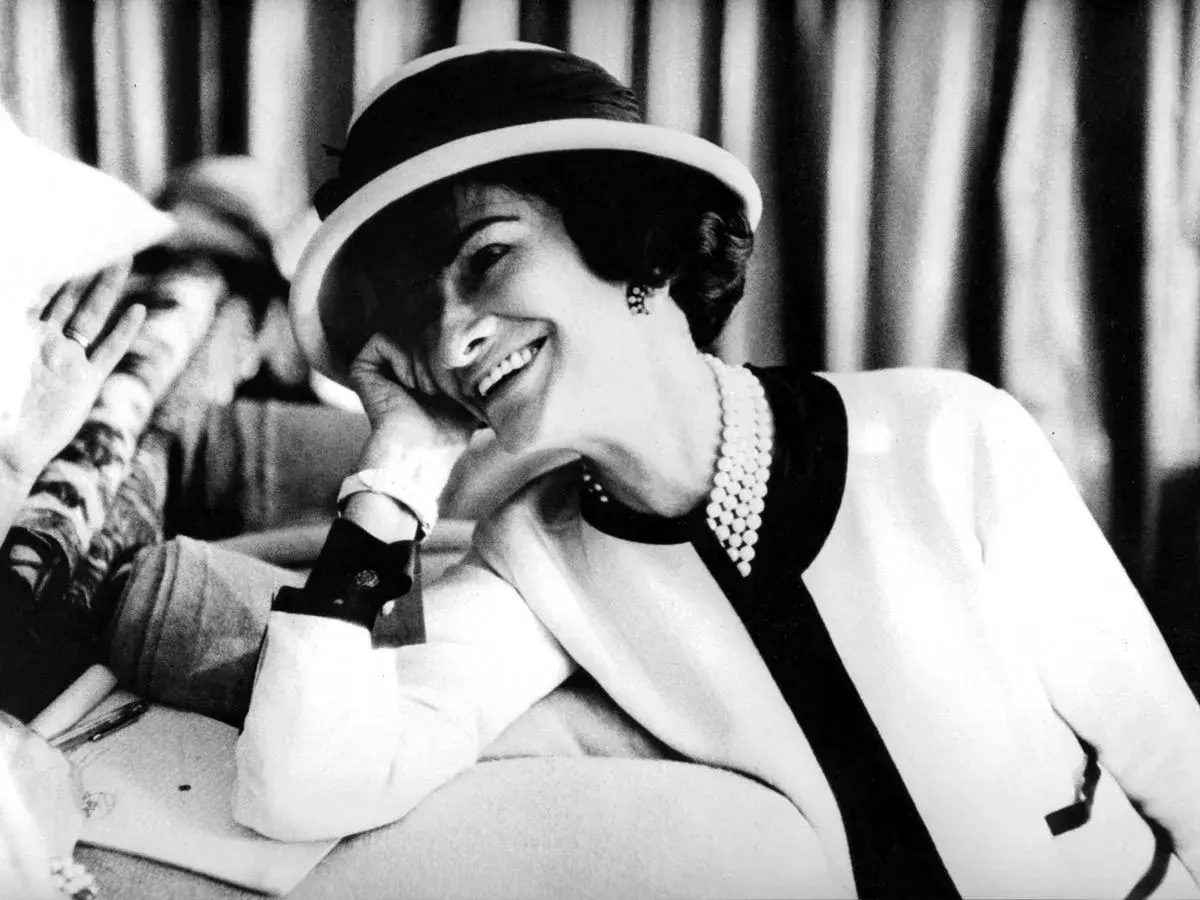 Coco Chanel, sonriendo con un sombrero y perlas, icónica figura de la moda.