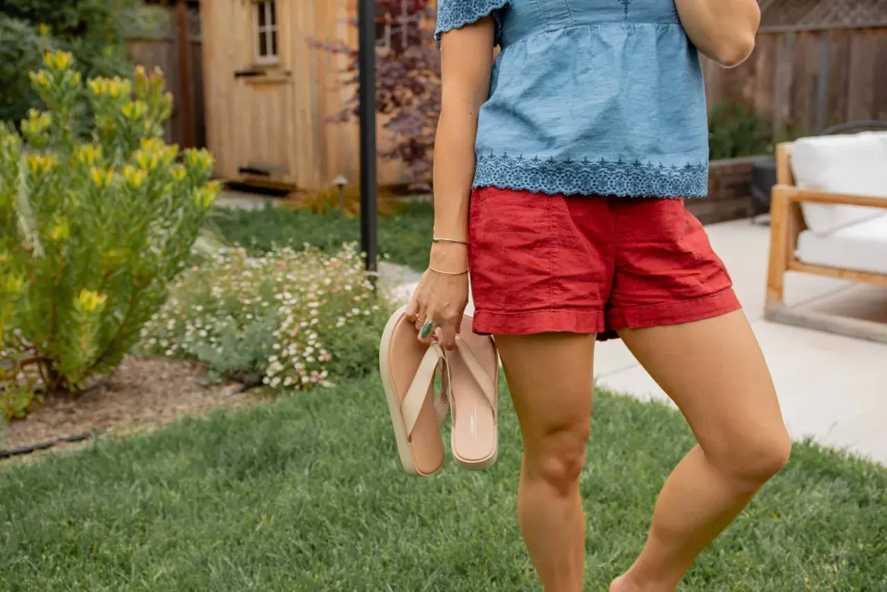 Mujer con blusa vaquera y shorts rojos, sosteniendo chanclas. El jardín tiene césped y flores.