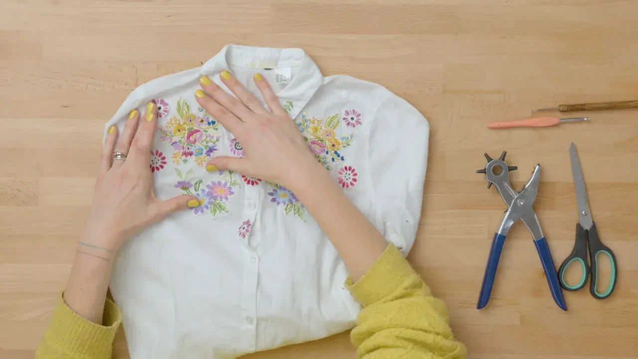 Manos embelleciendo una camisa blanca con bordados florales, lista para un proyecto de upcycling ropa. Herramientas de costura a un lado.