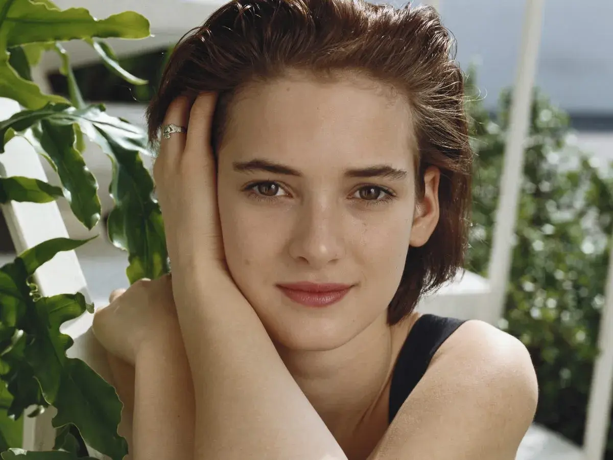 Winona Ryder joven, con su mano en la mejilla y un anillo discreto, rodeada de vegetación exuberante.