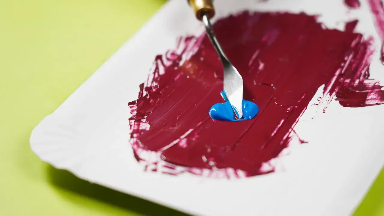 Mezclando azul con rojo para descubrir como se hace el color granate. Una espátula recoge pintura azul sobre una base roja.