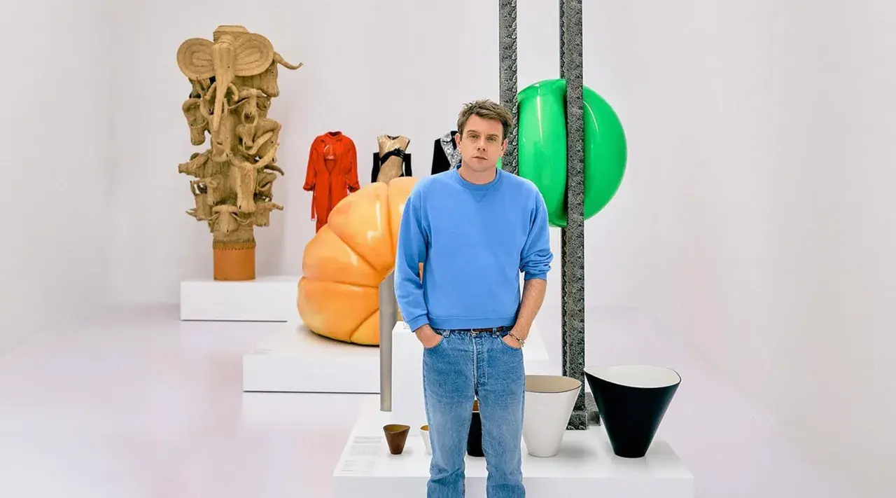 Jonathan Anderson posa entre esculturas y objetos de diseño, incluyendo una figura de madera tallada y un gran globo verde.