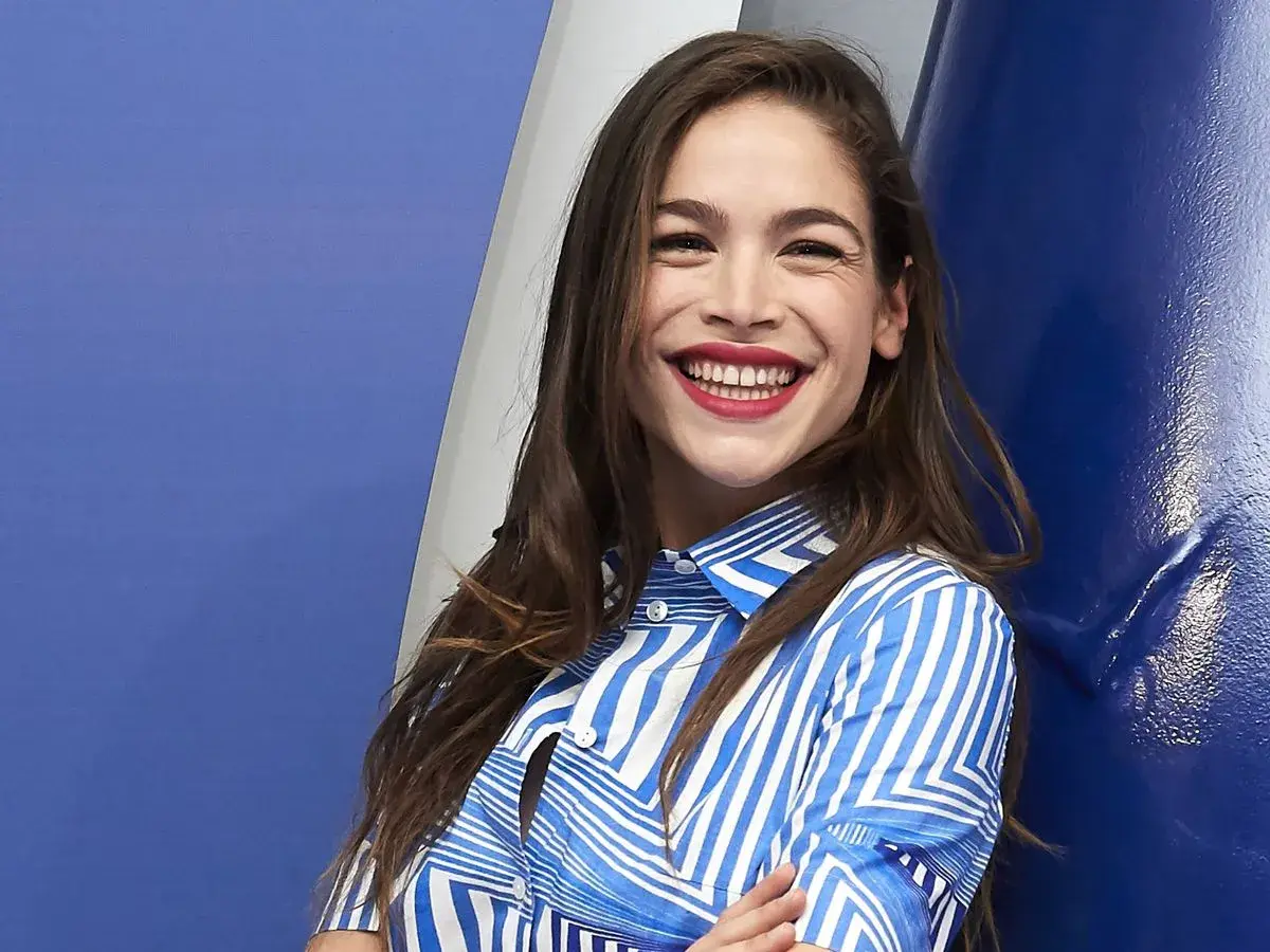 Cosima Ruiz de la Prada, sonriendo con labios rojos y camisa a rayas azules y blancas, se apoya en una pared azul.