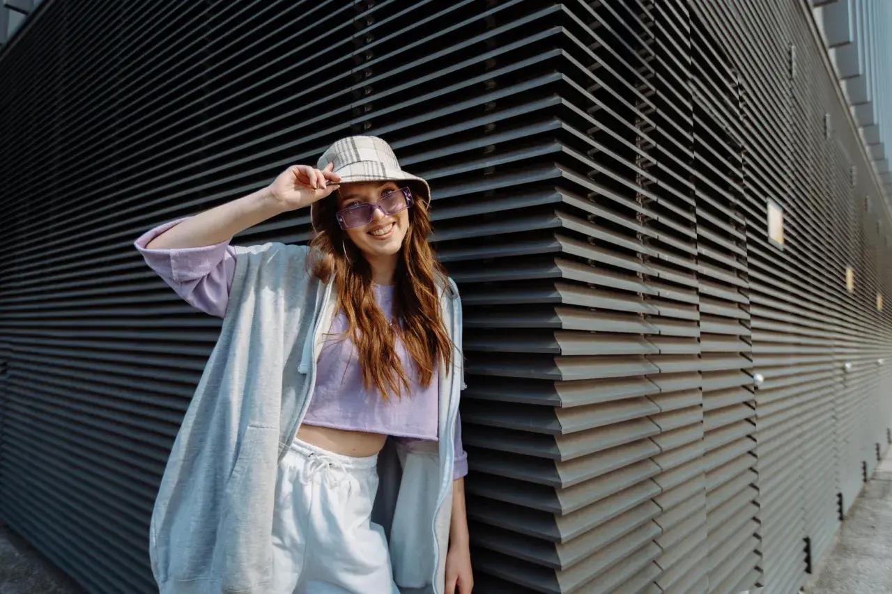 Chica vsco con sombrero y gafas de sol moradas, sonriendo y apoyada en una pared de lamas grises.