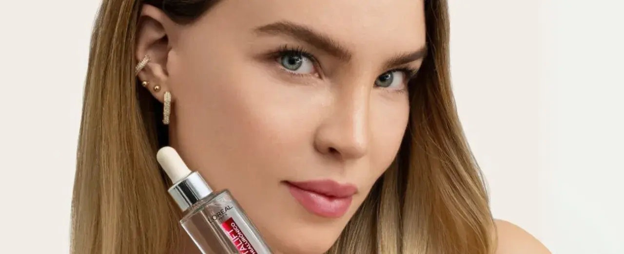 Mujer con cabello rubio y joyas brillantes sostiene un frasco de sérum, parte de su rutina de cuidado de la piel.