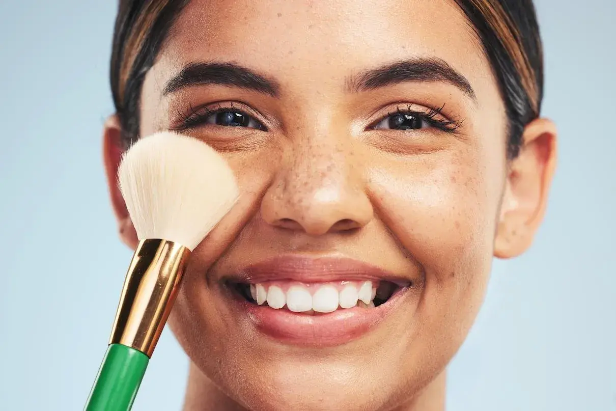 Una mujer sonríe mientras se aplica maquillaje con una brocha. Descubre trucos de maquillaje profesional para realzar tu belleza natural.