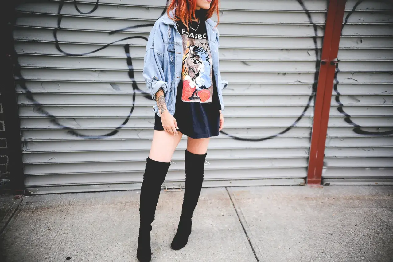 Chica con botas altas para bajitas, chaqueta vaquera y camiseta gráfica, posando frente a una pared de garaje con grafitis.