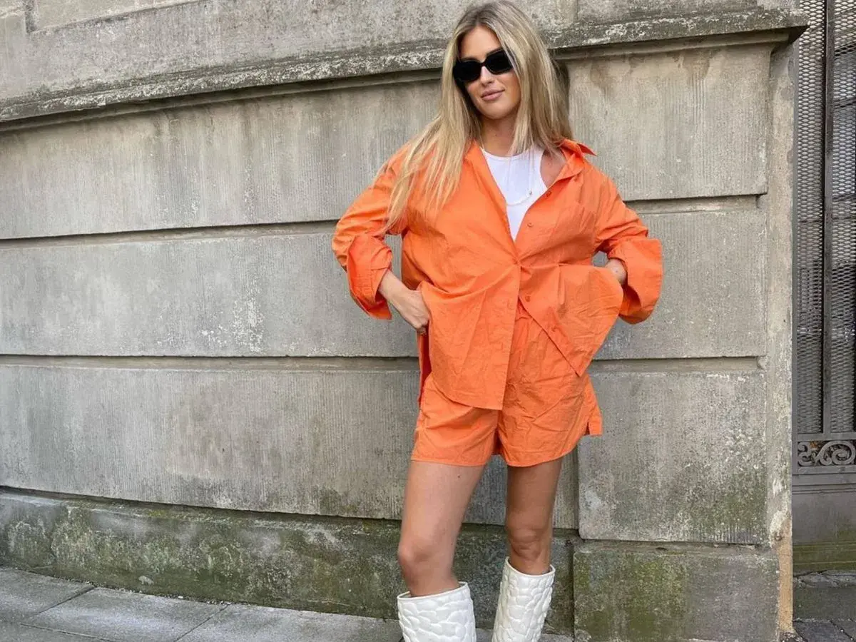 Mujer con conjunto naranja, gafas de sol y botas blancas. Se ve muy comfy y lista para pasear.