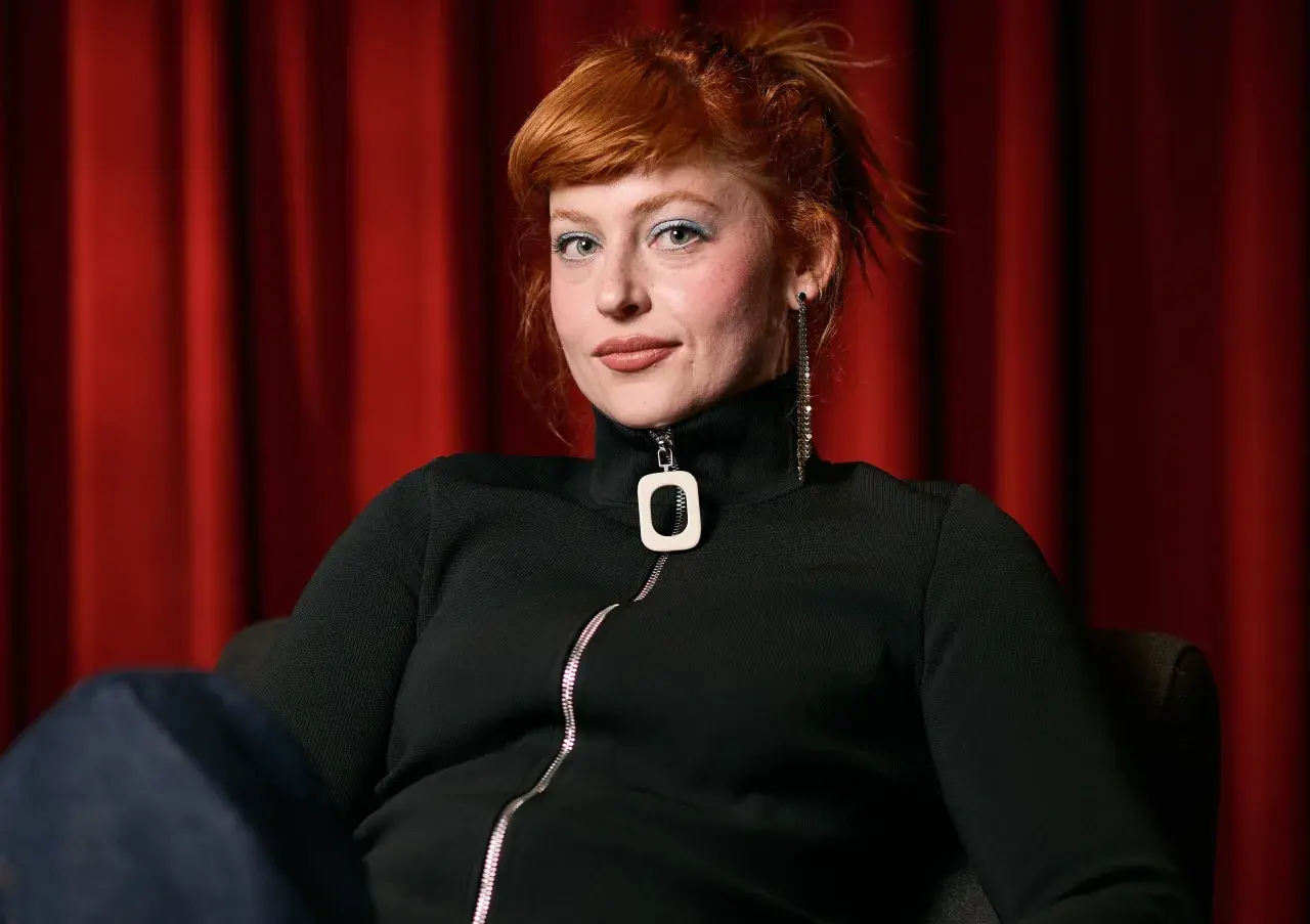 Chloe Wallace, con su cabello pelirrojo recogido y maquillaje azul, posa frente a una cortina roja.