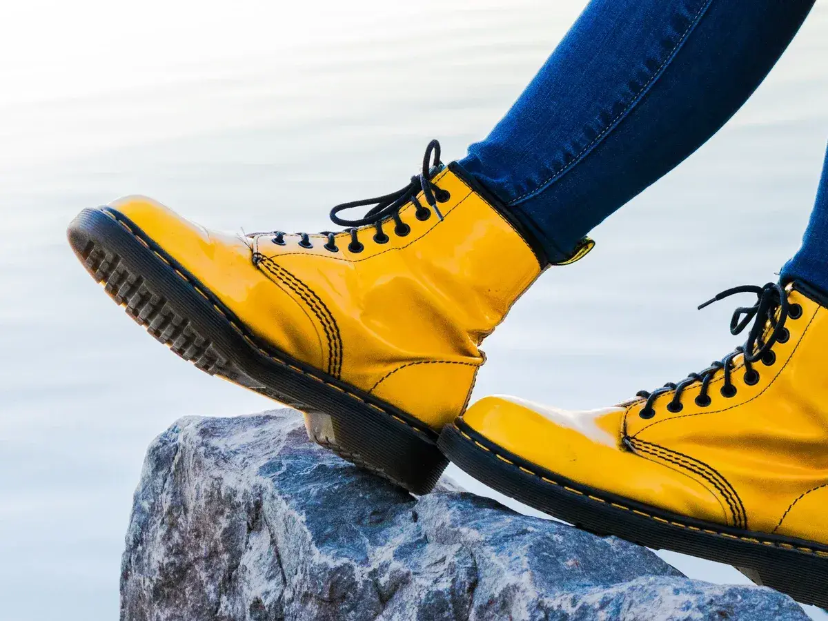Botas amarillas de charol, con cordones negros, pisan una roca. Las botas de piel ceden ante la aventura.