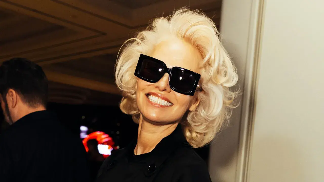 Pamela Anderson, con su icónica melena rubia y gafas de sol XL, sonríe con confianza.