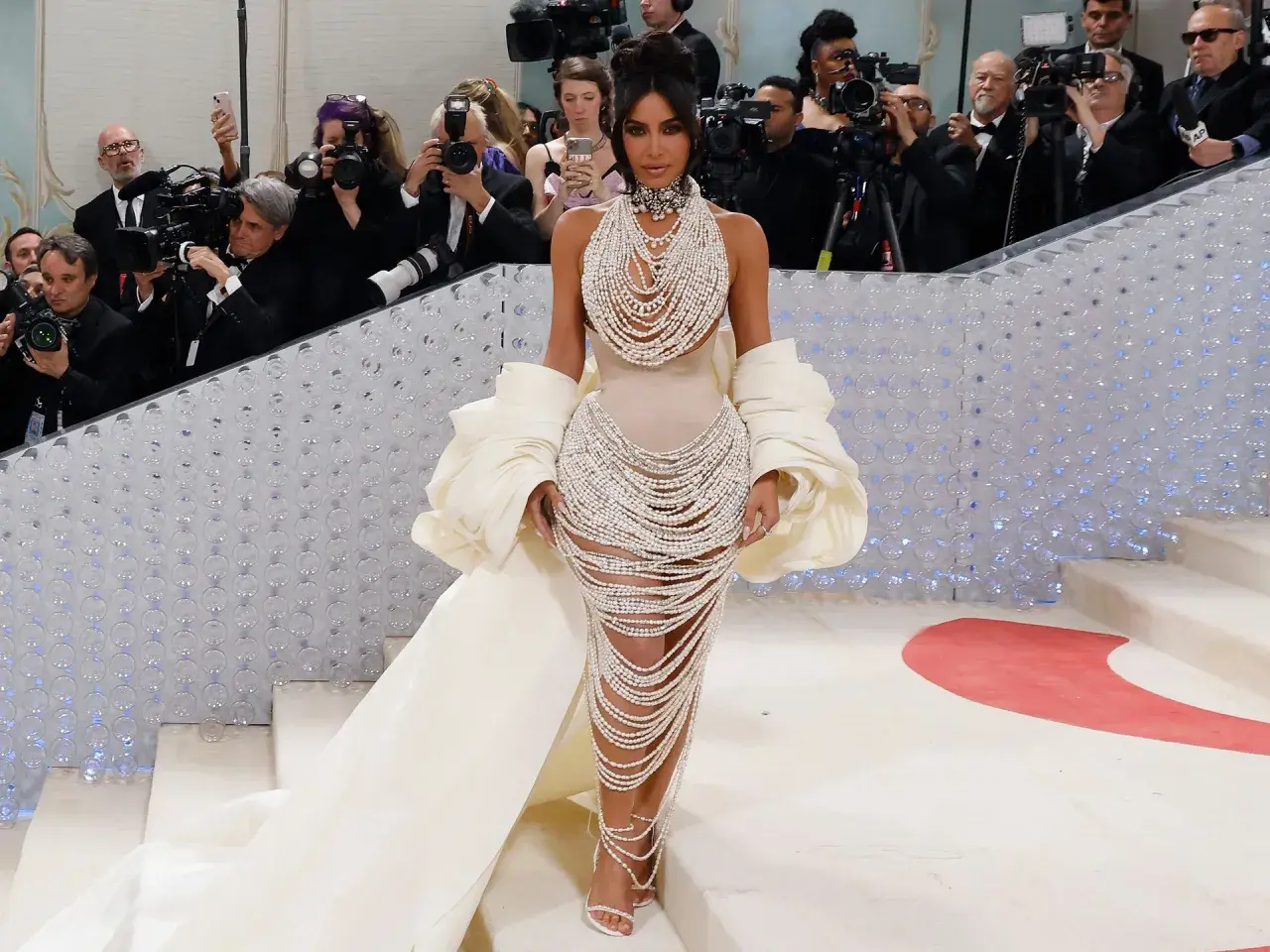 Kim Kardashian desciende la escalera de la Met Gala, deslumbrando con un vestido de perlas y una capa voluminosa, rodeada de fotógrafos.