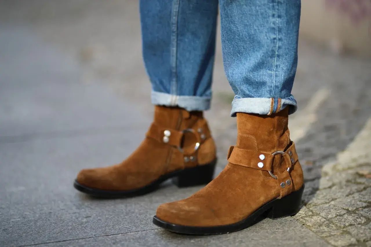 Botas de gamuza marrón con hebillas y jeans doblados. La piel girada de las botas contrasta con el denim.