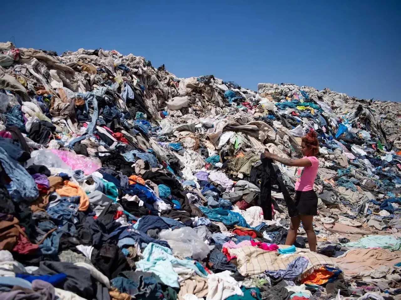 Montaña de ropa desechada, reflejo de las **fast fashion consecuencias**. Una joven busca entre los residuos textiles bajo un cielo azul.