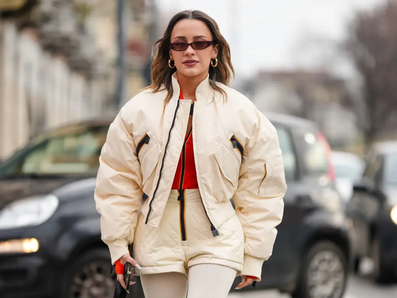 Mujer con gafas de sol y pendientes dorados, luciendo una bomber acolchada color crema sobre un top naranja y shorts a juego.