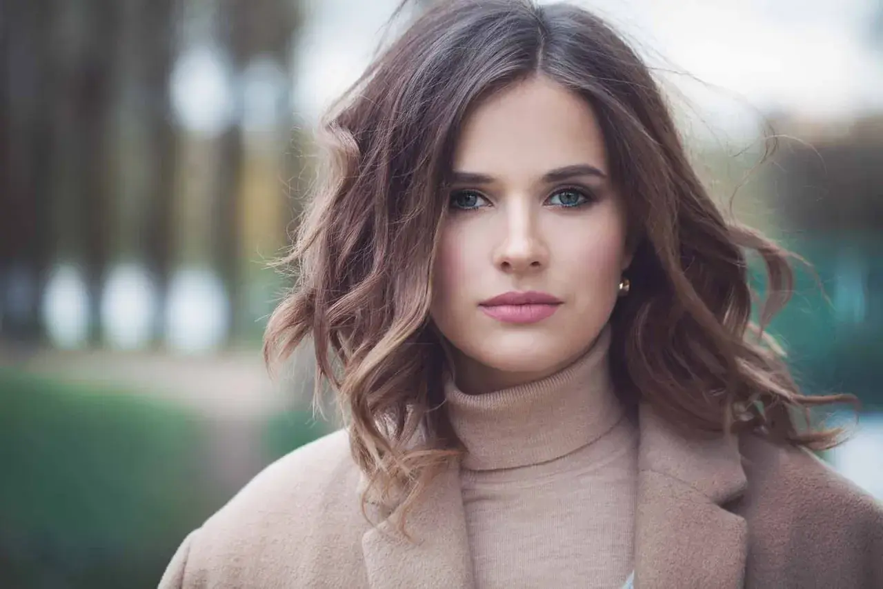 Mujer con una elegante media melena ondulada, ojos verdes y labios rosados, vestida con un abrigo beige.