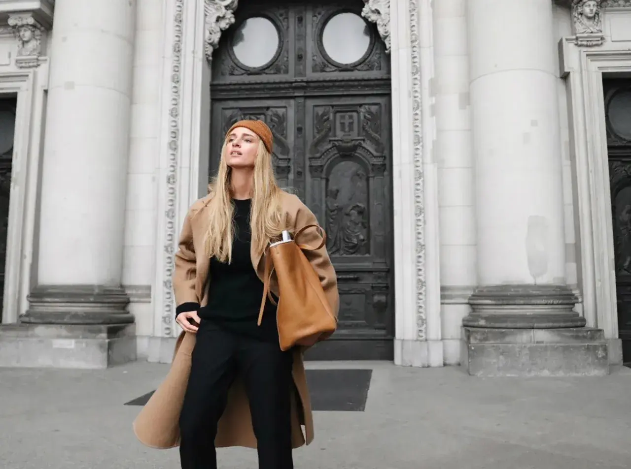 Mujer con abrigo camel, gorro y bolso marrón, luciendo un elegante pantalon negro.