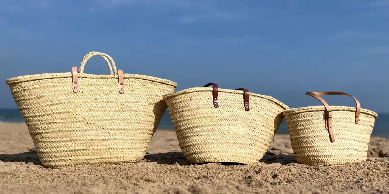 Tres capazos de playa grandes de palma, con asas de cuero, descansan en la arena bajo un cielo azul.