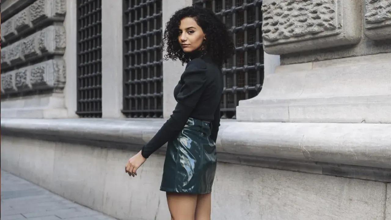 Joven con pelo rizado y falda verde brillante, estilo inspirado en Mary Quant, posa frente a un edificio histórico.