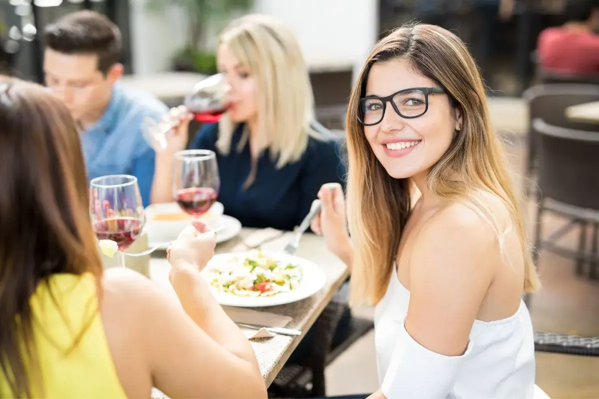 Chicas disfrutan una comida al mediodía, luciendo atuendos casuales y elegantes. La mujer con gafas sonríe a la cámara.